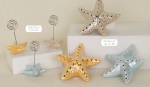 Stelle Marine in Porcellana Tema Mare 02A353 - 02A354 Una selezione di bomboniere a forma di stelle marine in porcellana pastello lucida poggia su supporti minimalisti bianchi. I modelli mostrati in 02A353-02A354.jpg includono una versione con clip metallica segnaposto e versioni più grandi con LED luminosi