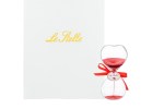 Clessidra in vetro con sabbia rossa e fiocco rosso decorativo per laurea, presentata accanto al suo astuccio bianco con scritta dorata Le Stelle.